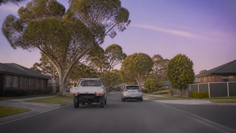 A work ute and a car driving along a quiet tree-lined Melbourne suburban street at dusk